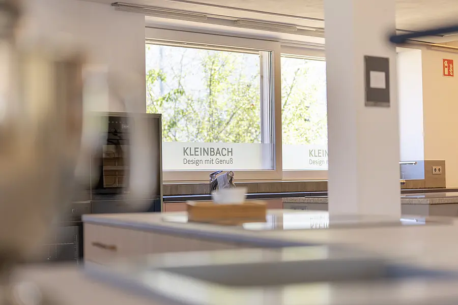 Blick durch die Küchenausstellung von Kleinbach mit Fenstern im Hintergrund mit der Aufschrift "KLEINBACH – Design mit Genuss". Man sieht grüne Bäume durch die Fenster.
